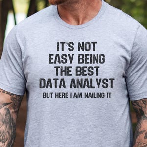 May include: A light gray short-sleeved t-shirt featuring black distressed-style text that reads: IT'S NOT EASY BEING THE BEST DATA ANALYST BUT HERE I AM NAILING IT. The shirt has a crew neckline and a heathered texture throughout the fabric.