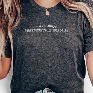 May include: Charcoal grey crew neck t-shirt with the white text "not bossy, aggressively helpful". A gold pendant necklace is visible. A tattoo on the arm reads "Mon Océan".