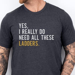 May include: Dark gray t-shirt with the text "YES, I REALLY DO NEED ALL THESE LADDERS." The word "LADDERS" is in gold. The shirt is a crew neck and has short sleeves.