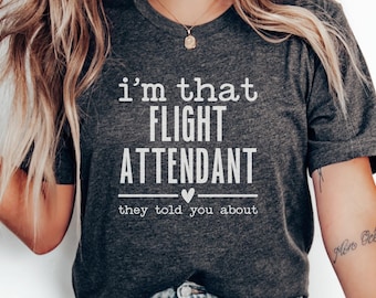 May include: Charcoal grey t-shirt featuring the white text "i'm that FLIGHT ATTENDANT they told you about". The design includes a heart graphic. The top is made of a soft fabric.