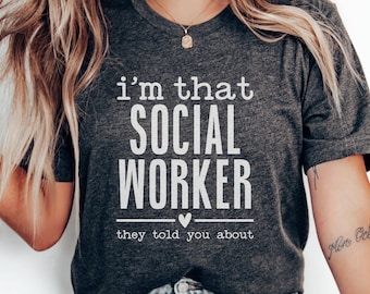 May include: Dark gray t-shirt with the text "i'm that SOCIAL WORKER they told you about" in white. The shirt has a crew neck and short sleeves. A small heart is below the word WORKER.