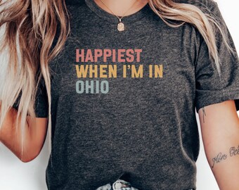 May include: Dark gray t-shirt with the text "Happiest When I'm in Ohio" in pink, yellow, and blue. The shirt is a crew neck and has short sleeves. The image shows the shirt on a person.