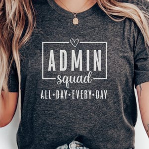 May include: Dark grey t-shirt featuring the text "ADMIN squad" in a white rectangular box, with a heart above "ADMIN". Below the box, the text reads "ALL • DAY • EVERY • DAY".