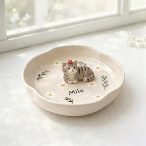 Op de afbeelding: Een beige keramische kattenbak met een geschulpte rand. De bak heeft een klein, grijs en wit kattenbeeldje met een rode hoed. De bak is versierd met witte madeliefjes en de naam "Milo".