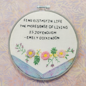 May include: Embroidered hoop art with a white background and a quote by Emily Dickinson: "Find ecstasy in life, the mere sense of living is joy enough." The quote is surrounded by a border of pink and purple flowers.