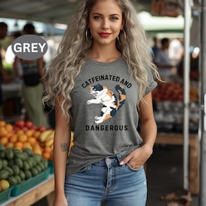 Op de afbeelding: Een grijs t-shirt met de tekst "Caffeinated and Dangerous" en een afbeelding van een lapjeskat. De kat is afgebeeld in een dynamische pose. Het t-shirt wordt gedragen met een blauwe jeans.