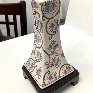 May include: A pale pink ceramic vase with a square base, adorned with pink floral patterns and gold accents. The vase sits on a dark brown wooden stand, showcasing an elegant, vintage design. The floral pattern includes pink roses and green leaves.