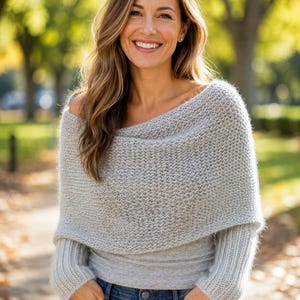 May include: A light gray knit sweater featuring a wide, folded-over cowl neckline that drapes over the shoulders, long sleeves with ribbed cuffs, and a textured, fine-gauge knit pattern, paired with classic blue denim jeans.