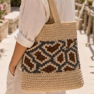 Op de afbeelding: Een beige gehaakte tote bag met een opvallend geometrisch patroon in donkerbruin en antracietgrijs. De tas is volledig gemaakt met een gestructureerde knoopsteek en is voorzien van twee bijpassende schouderbanden om de tas te dragen.