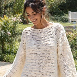 May include: An off-white, long-sleeved crochet tunic featuring a wide boat neckline and a textured, horizontal stitch pattern. The garment has a loose, relaxed fit with wide sleeves that extend to the wrists, crafted from a thick, soft yarn.