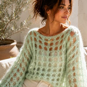 Puede incluir: Un suéter verde claro de tejido abierto con mangas largas y voluminosas. El suéter se combina con pantalones de color claro. La modelo está sentada en un sofá de tonos neutros, con una planta en el fondo.
