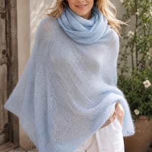May include: A light blue knit poncho with a matching scarf. The poncho has a loose, flowing design and is paired with white pants. The outfit is soft and airy, perfect for layering. The model is smiling.