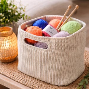May include: A cream-colored, rectangular, crocheted storage basket with two integrated side handles. The basket is filled with several colorful balls of yarn in shades of blue, orange, green, and purple, along with two wooden knitting needles.