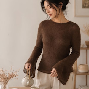 May include: A brown long-sleeved sweater with flared sleeves. The model is wearing white pants and holding a small teapot. The image has the text "COZY. CHIC. UNIQUELY YOU."