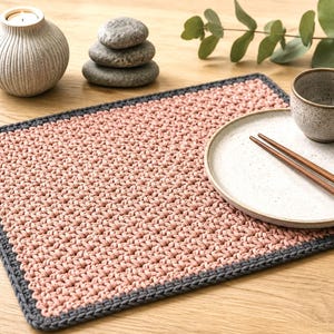 May include: A rectangular crochet placemat featuring a textured dusty pink center and a solid dark gray border. The placemat is crafted with a dense, uniform stitch pattern, providing a structured surface for dining items like plates and bowls.