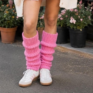 May include: Bright pink knitted leg warmers are worn over white trainers with pink stripes. The leg warmers are scrunched down, creating a layered effect. The trainers have a brown sole. The leg warmers appear to be made of wool.