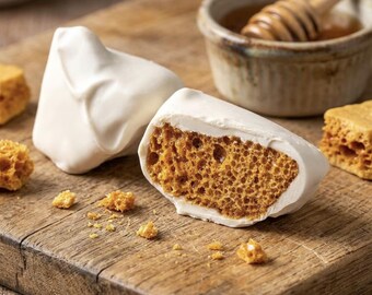 May include: Two pieces of honeycomb candy partially coated in a smooth white chocolate layer. The interior of the candy reveals a porous, golden-brown, aerated texture. Small golden crumbs are scattered nearby, alongside a small bowl of honey.
