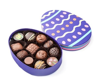 May include: An open, purple, egg-shaped box of assorted chocolates. The chocolates are various shapes and sizes, with different toppings and designs. The lid, also egg-shaped, is purple with a pattern of wavy lines and dots. A festive treat.
