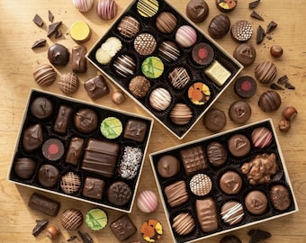 May include: Three rectangular boxes filled with an assortment of dark, milk, and white chocolate truffles and pralines. The chocolates feature various shapes, textures, and patterns, including stripes, swirls, and printed designs, scattered on a wooden surface.