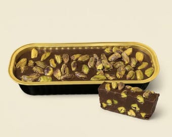 May include: A rectangular black and gold container filled with dark chocolate fudge topped with whole green pistachios. A single rectangular piece of the same dark chocolate and pistachio fudge sits in front of the container, showing the interior texture.