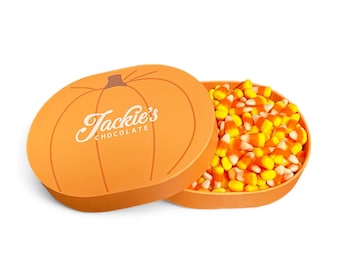May include: An orange pumpkin-shaped box with the text "Jackie's Chocolate" in a cursive font. The box is open, revealing a container filled with candy corn in yellow, orange, and white colors. The box is on a white background.