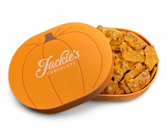 May include: An open orange pumpkin-shaped box filled with peanut brittle. The box lid features a brown stem and the text "Jackie's Chocolate" in a cursive font. The brittle pieces are golden brown and irregularly shaped.