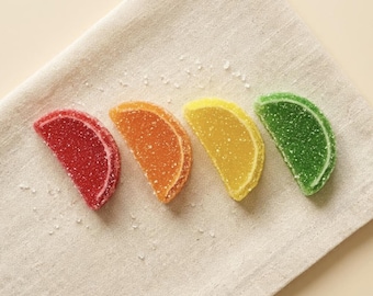May include: Four crescent-shaped fruit-flavored jelly candies coated in granulated sugar. The candies are arranged in a row, featuring red, orange, yellow, and green colors, each with a distinct rind-like edge and a translucent, textured surface.
