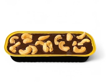 May include: A rectangular dessert in a gold-colored container. The dessert has a dark brown, glossy topping, and is garnished with several whole cashews. The container has a black base.