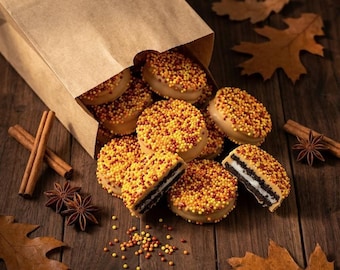 May include: Round sandwich cookies coated in tan icing and topped with small yellow, orange, and brown sprinkles spilling from a brown paper bag. The cookies feature a dark chocolate center with white cream filling, accompanied by cinnamon sticks and star anise.