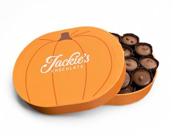 May include: A round orange box shaped like a pumpkin with the text Jackie's CHOCOLATE printed in white on the lid. The lid is partially open, revealing a collection of dark brown round chocolates nestled inside individual paper liners.