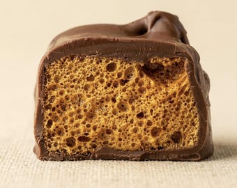 May include: A single piece of chocolate-covered honeycomb candy, cut in half to reveal a porous, golden-brown interior texture. The exterior is coated in a smooth, solid milk chocolate layer that wraps around the brittle, airy center.