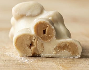 May include: A piece of white chocolate candy with visible whole roasted nuts embedded inside. The confection has a smooth, creamy white exterior coating that contrasts with the tan, textured appearance of the nuts exposed on the cut surface.