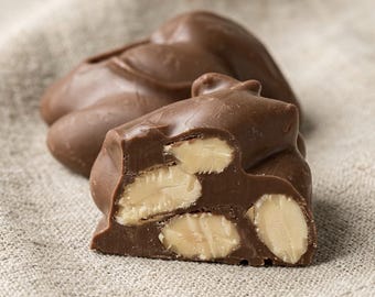 May include: A milk chocolate cluster with visible whole almonds embedded inside. One piece is broken in half to reveal the light-colored nut pieces encased within the smooth, brown chocolate coating, while another whole piece sits behind it.