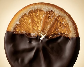 May include: A single circular slice of dried orange, with the bottom half coated in a smooth, dark brown chocolate layer. The top half reveals the translucent, fibrous, and golden-orange pulp structure of the fruit, contrasting with the dark coating.