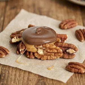 May include: A cluster of brown pecan halves topped with a layer of golden caramel and a smooth, circular dollop of milk chocolate. The treat is arranged on a piece of light tan parchment paper, with additional loose pecans scattered nearby.