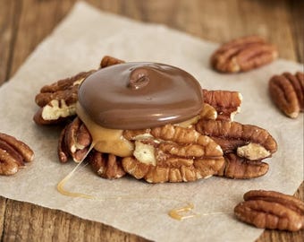 May include: A cluster of brown pecan halves topped with a layer of golden caramel and a smooth, circular dollop of milk chocolate. The treat is arranged on a piece of light tan parchment paper, with additional loose pecans scattered nearby.