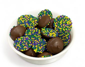 May include: A white bowl filled with chocolate-covered candies. The candies are round and dark brown, topped with colorful sprinkles in yellow, green, and purple. The bowl is set against a white background.