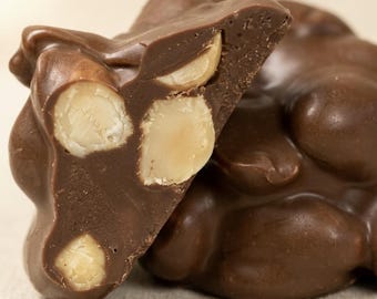 May include: A cluster of milk chocolate candy with a broken piece in the foreground revealing several whole macadamia nuts embedded inside the smooth, brown chocolate. The texture of the chocolate is solid and glossy with visible nut pieces.