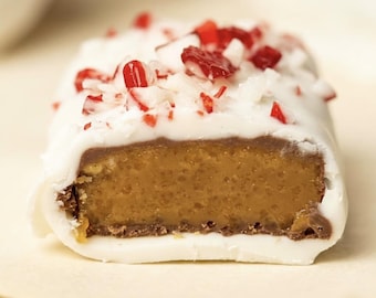 May include: A rectangular candy bar coated in white chocolate with a smooth, light brown caramel-colored center. The top is garnished with small, crushed pieces of red and white peppermint candy, and a thin layer of milk chocolate is visible.