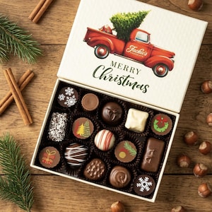 May include: A square white gift box filled with assorted chocolates, featuring a red truck carrying a Christmas tree and the text Jackie's Chocolate Merry Christmas on the lid. The chocolates have various shapes, colors, and holiday designs.