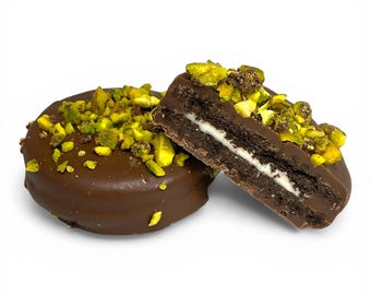 May include: Two chocolate-covered sandwich cookies, one whole and one cut in half, with a white cream filling. The cookies are topped with chopped green pistachios. The image is set against a white background.