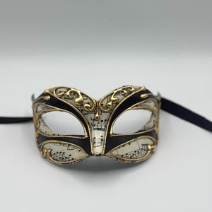 May include: A black and gold masquerade mask with a white musical note pattern. The mask has a black ribbon attached.