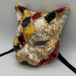 May include: A decorative cat-shaped mask with a harlequin pattern in black, white, red, and gold. The mask features musical notes and gold filigree accents. Black ribbons are attached for wearing.