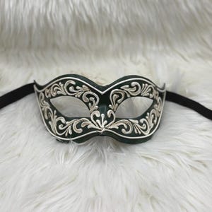 Op de afbeelding: Een donkergroen masker voor een gemaskerd bal met ingewikkeld wit krulwerk. Het masker heeft oogopeningen en een zwarte elastische band. Het ontwerp is symmetrisch en elegant, geschikt voor een formeel evenement of kostuum.