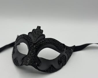 Può includere: Una maschera nera da ballo in maschera con un design ornato. La maschera presenta aperture per gli occhi ed è collegata a un nastro nero. La maschera ha una superficie strutturata e un elemento decorativo a forma di corona nella parte superiore.