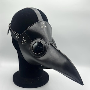 Peut inclure: Un masque de médecin de la peste en cuir noir avec un long bec incurvé et de grandes ouvertures rondes pour les yeux. Le masque a une sangle qui passe sur la tête et est fixée avec des rivets. Le masque est exposé sur une forme de tête noire.