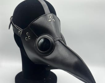 May include: A black leather plague doctor mask with a long, curved beak and large, round eye openings. The mask has a strap that goes over the head and is secured with rivets. The mask is displayed on a black head form.