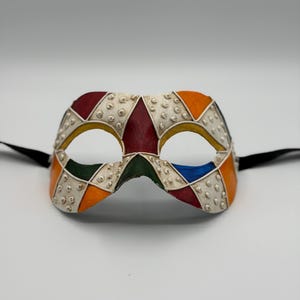 以下が含まれることがあります： 幾何学模様のカラフルな仮面舞踏会のマスク。マスクは、盛り上がった銀色のドットが付いた白いベースで、赤、オレンジ、黄色、緑、青のひし形のセグメントで飾られています。黒いリボンが両側から伸びています。