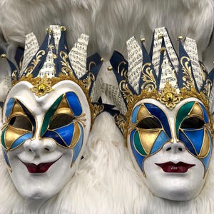 May include: Two Venetian-style masks with a white base, featuring blue, green, and gold geometric designs. The masks have a smiling face and a jester-style crown with musical notes. The masks are set against a white, fluffy background.