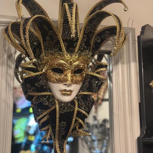 Peut inclure: Un masque de carnaval vénitien noir et or avec un chapeau de bouffon. Le masque a un visage blanc avec des accents dorés et des yeux noirs. Le chapeau est en velours noir avec des garnitures dorées et a de longues cornes pointues.